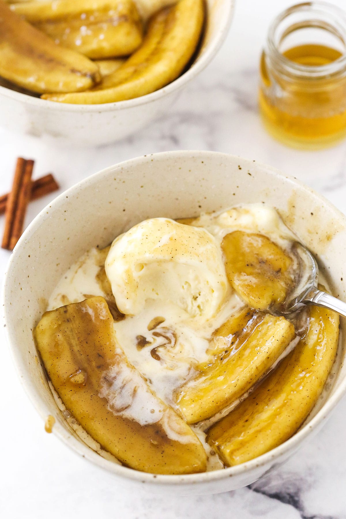 A bowl full of bananas foster and ice cream on top of a marble counter with cinnamon sticks and more bananas beside it