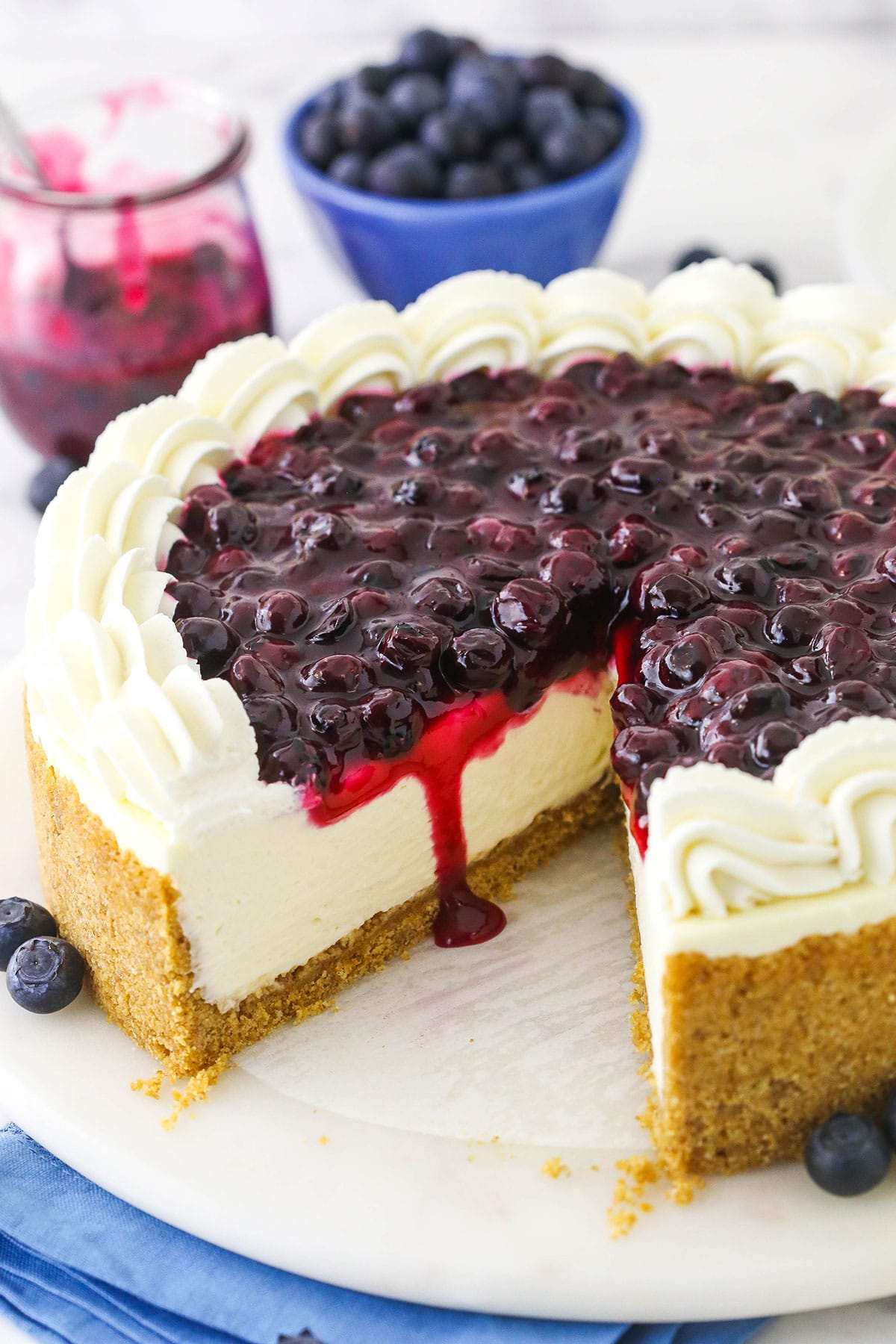 A blueberry cheesecake with a graham cracker crust on a serving platter with one large slice removed from the cake