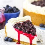 A piece of blueberry cheesecake on a plate with a bowl of blueberries and the remaining cake behind it