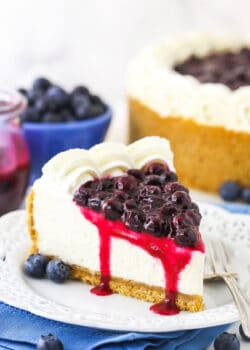 A piece of blueberry cheesecake on a plate with a bowl of blueberries and the remaining cake behind it