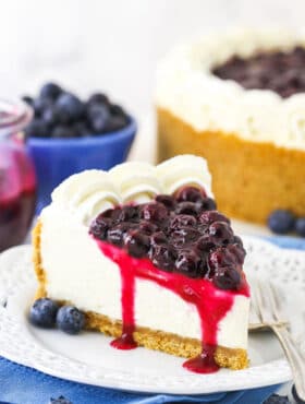 A piece of blueberry cheesecake on a plate with a bowl of blueberries and the remaining cake behind it