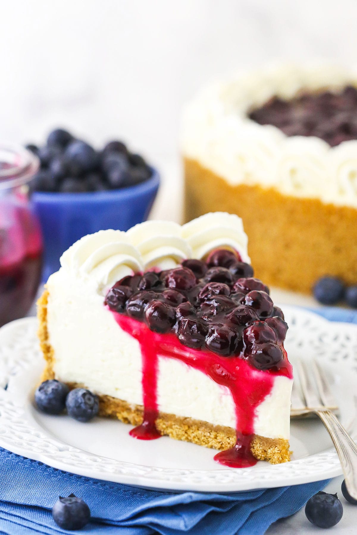 A piece of blueberry cheesecake on a plate with a bowl of blueberries and the remaining cake behind it