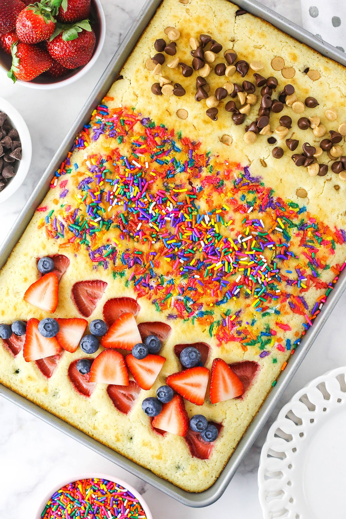 A sheet pan full of cooked pancakes with fruit, sprinkles and chocolate & peanut butter chips on top