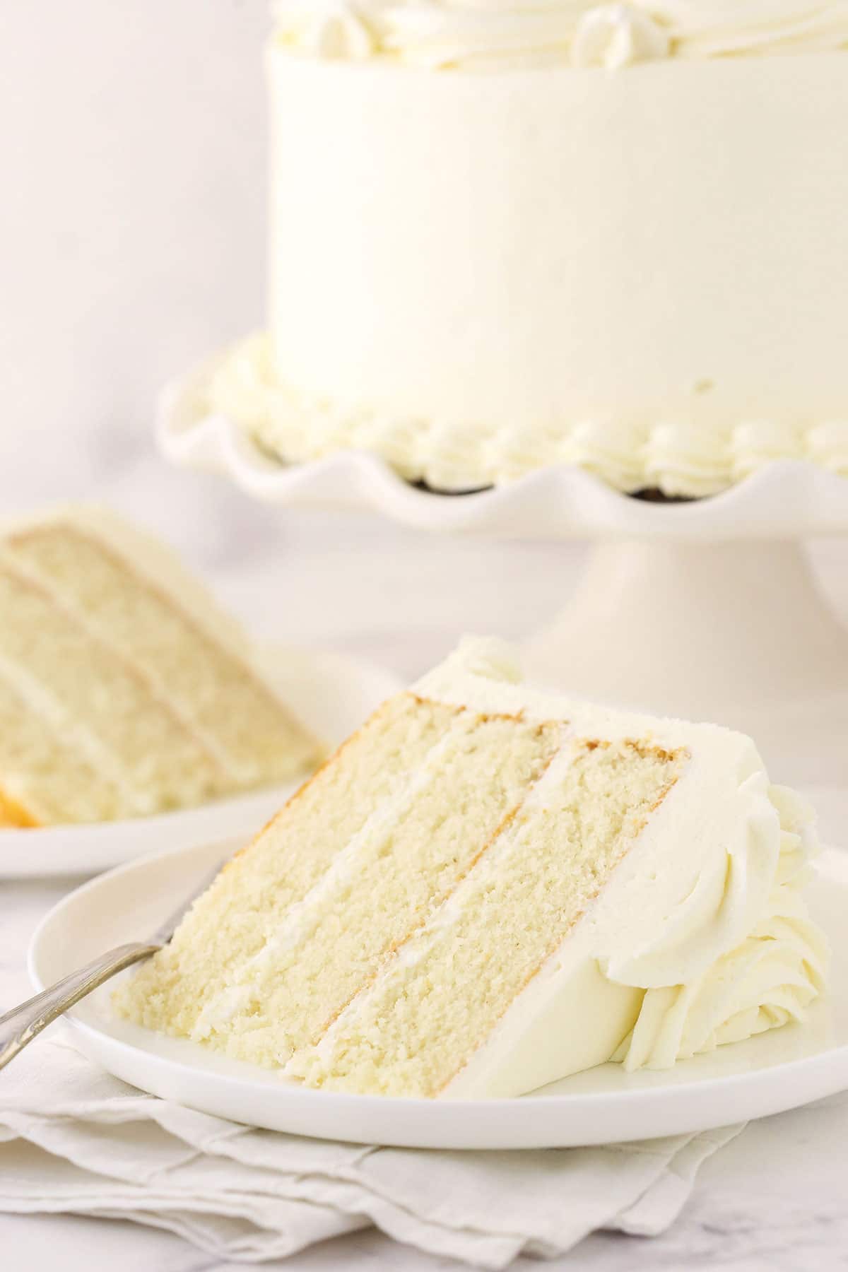 A big slice of vanilla layer cake on a plate with the full cake on a cake stand in the background