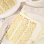 A piece of white cake on a plate with another slice and the remaining cake behind it