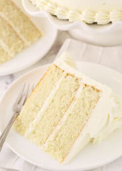 A piece of white cake on a plate with another slice and the remaining cake behind it