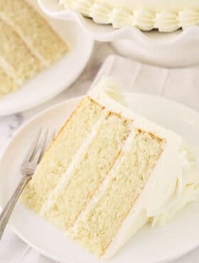 A piece of white cake on a plate with another slice and the remaining cake behind it