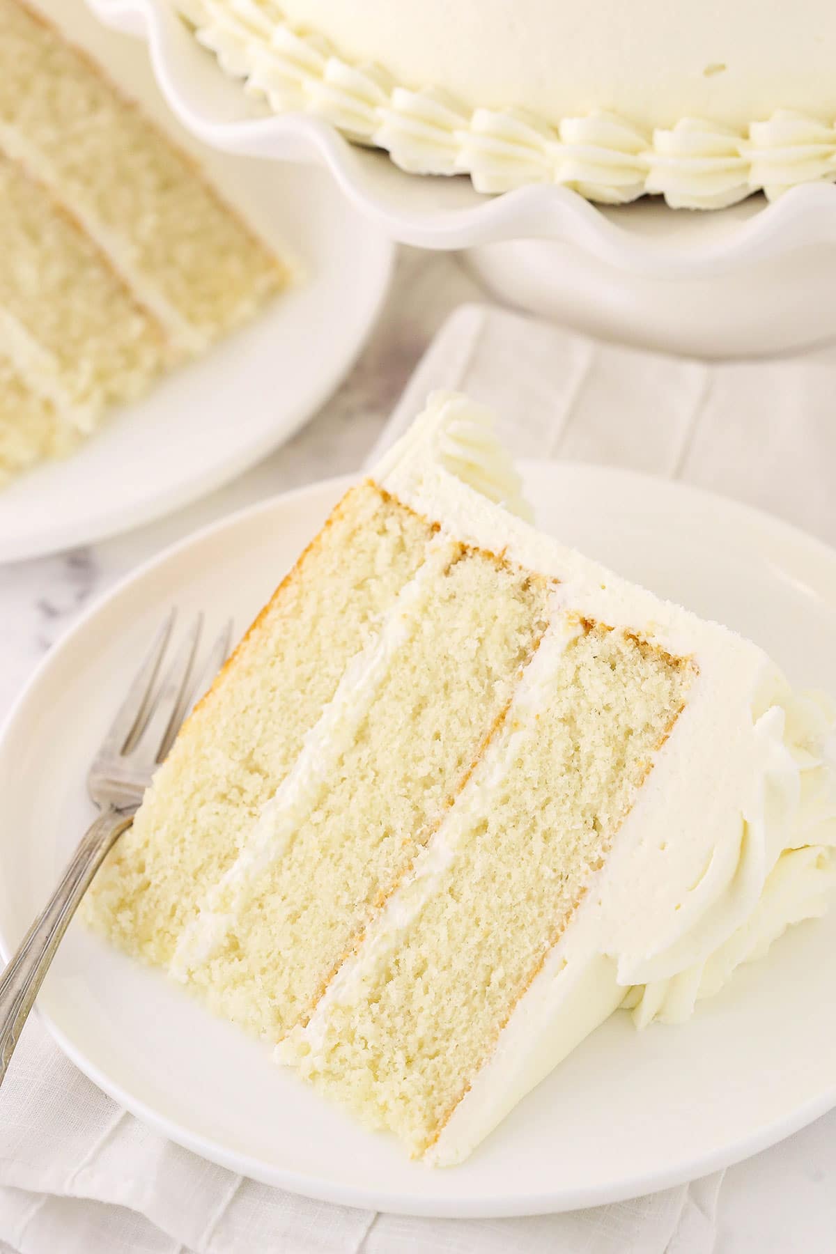 A piece of white cake on a plate with another slice and the remaining cake behind it