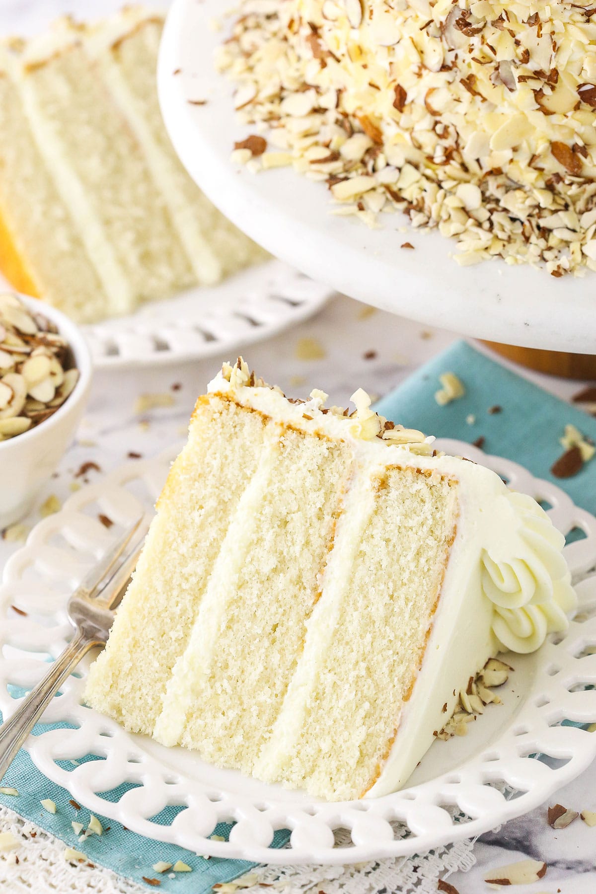 slice of white almond cream cake on white plate and blue napkin