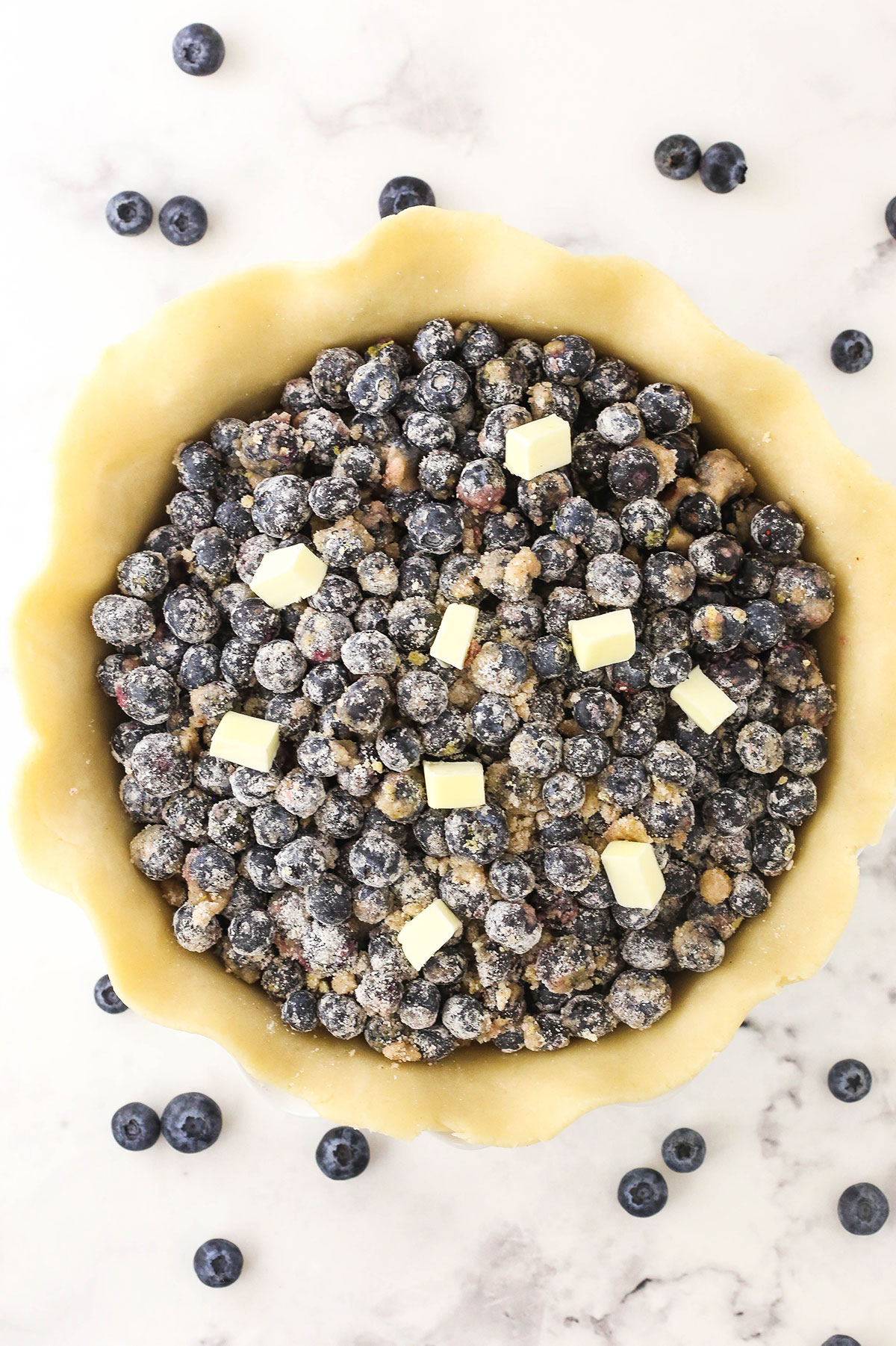 An unbaked pie crust in a pan with blueberry filling inside and small cubes of butter on top of the filling
