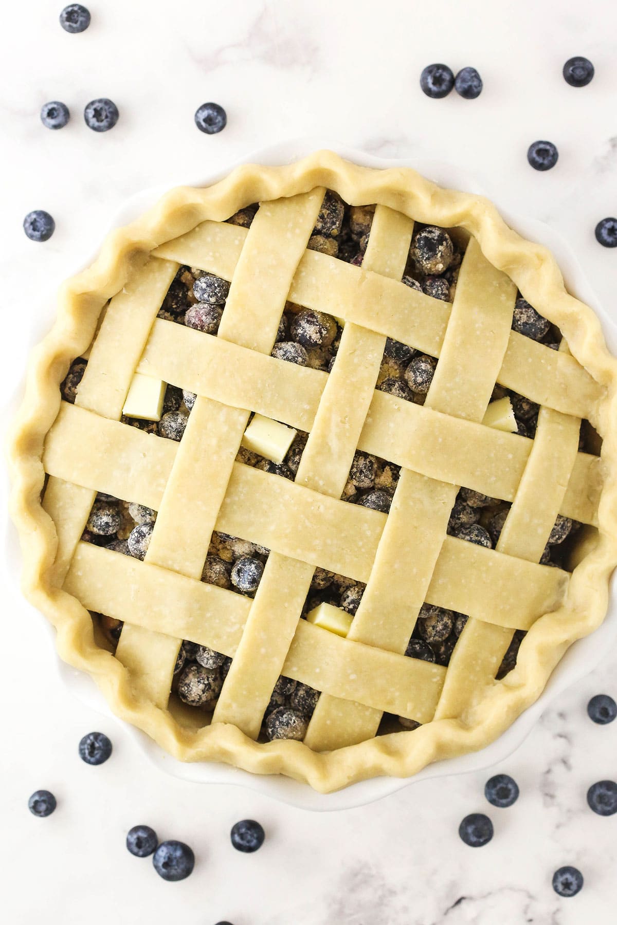 An assembled, unbaked blueberry pie on a marble countertop with blueberries scattered about