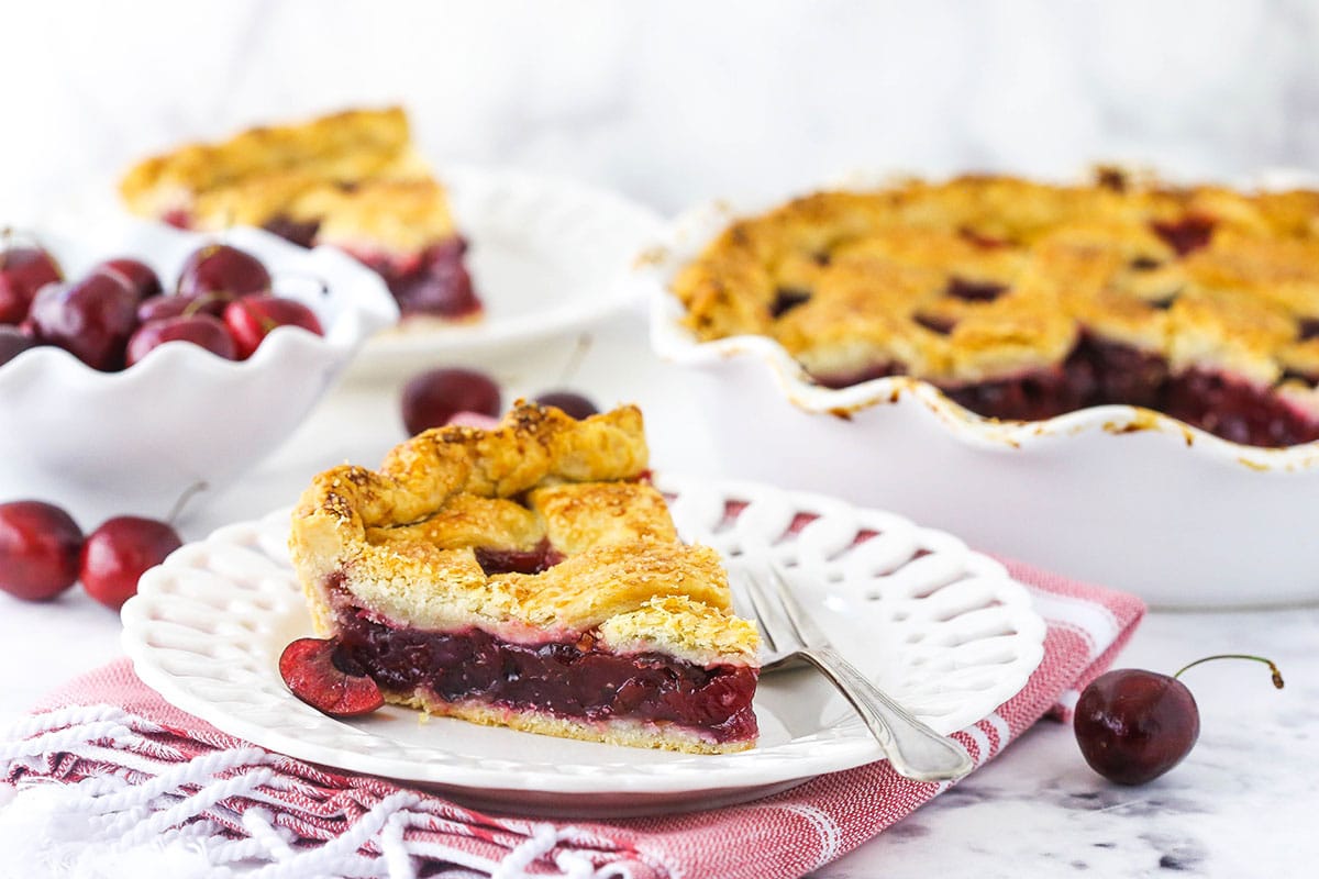 A slice of cherry pie on a plate with another slice and the remaining pie behind it