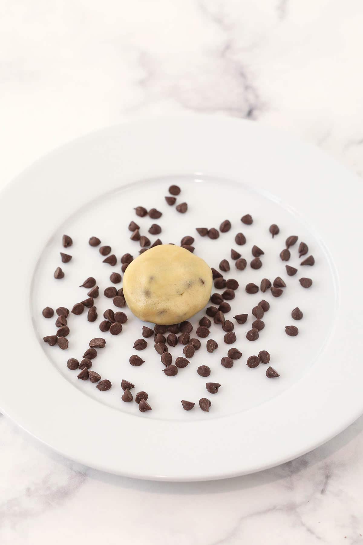 A ball of chocolate chip cookie dough being pressed into a few extra chocolate chips on a small plate
