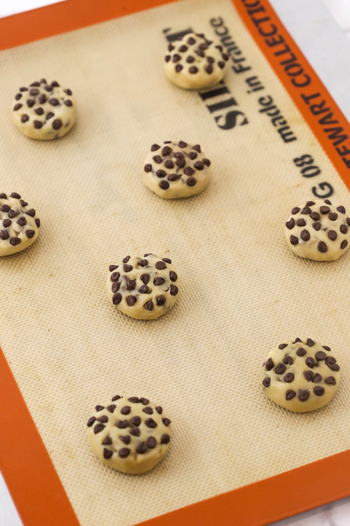 Eight balls of chocolate chip cookie dough on a lined cookie sheet with extra mini chocolate chips pressed into each one
