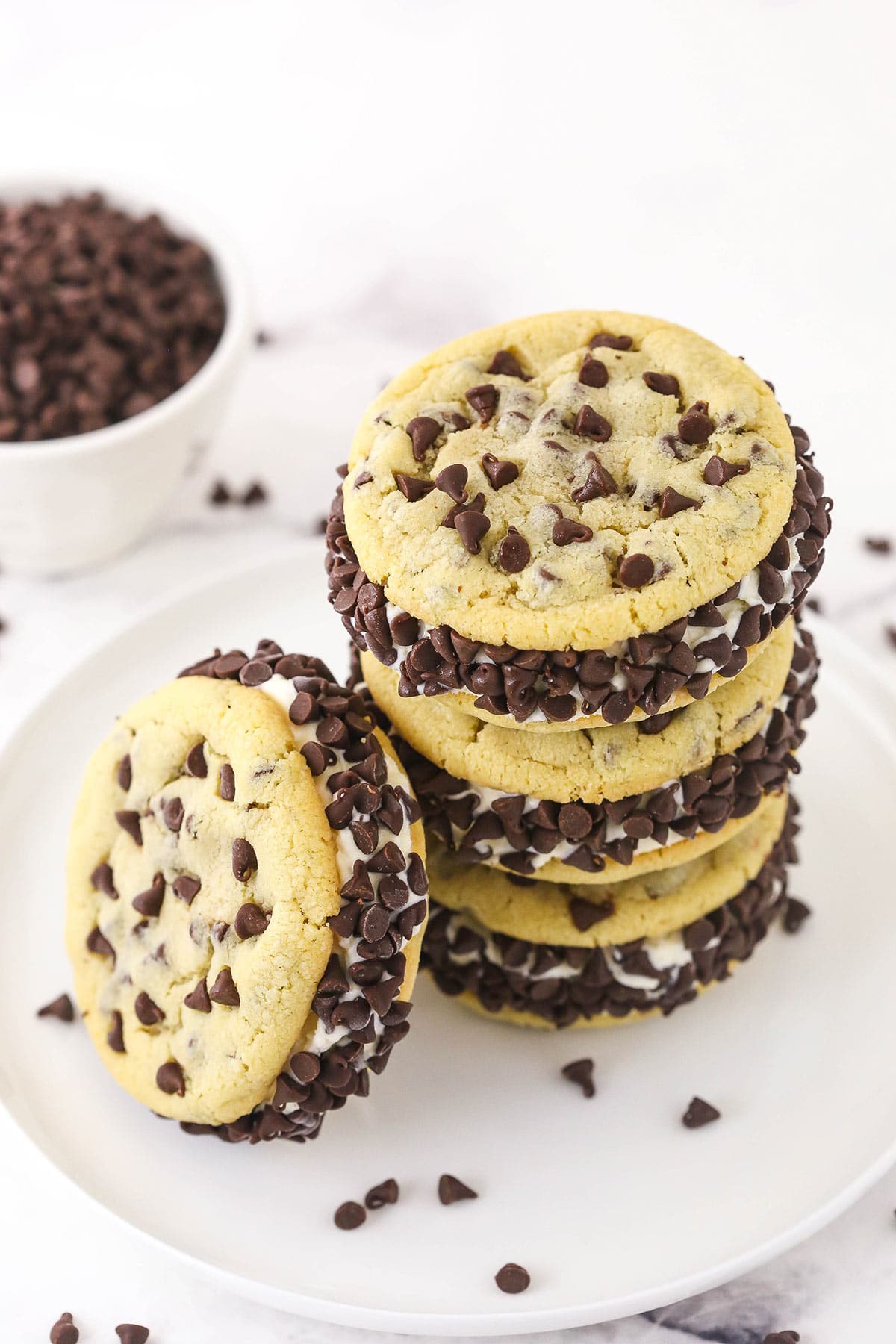 Four chocolate chip cookie ice cream sandwiches on a plate with a bowl of mini chocolate chips in the background