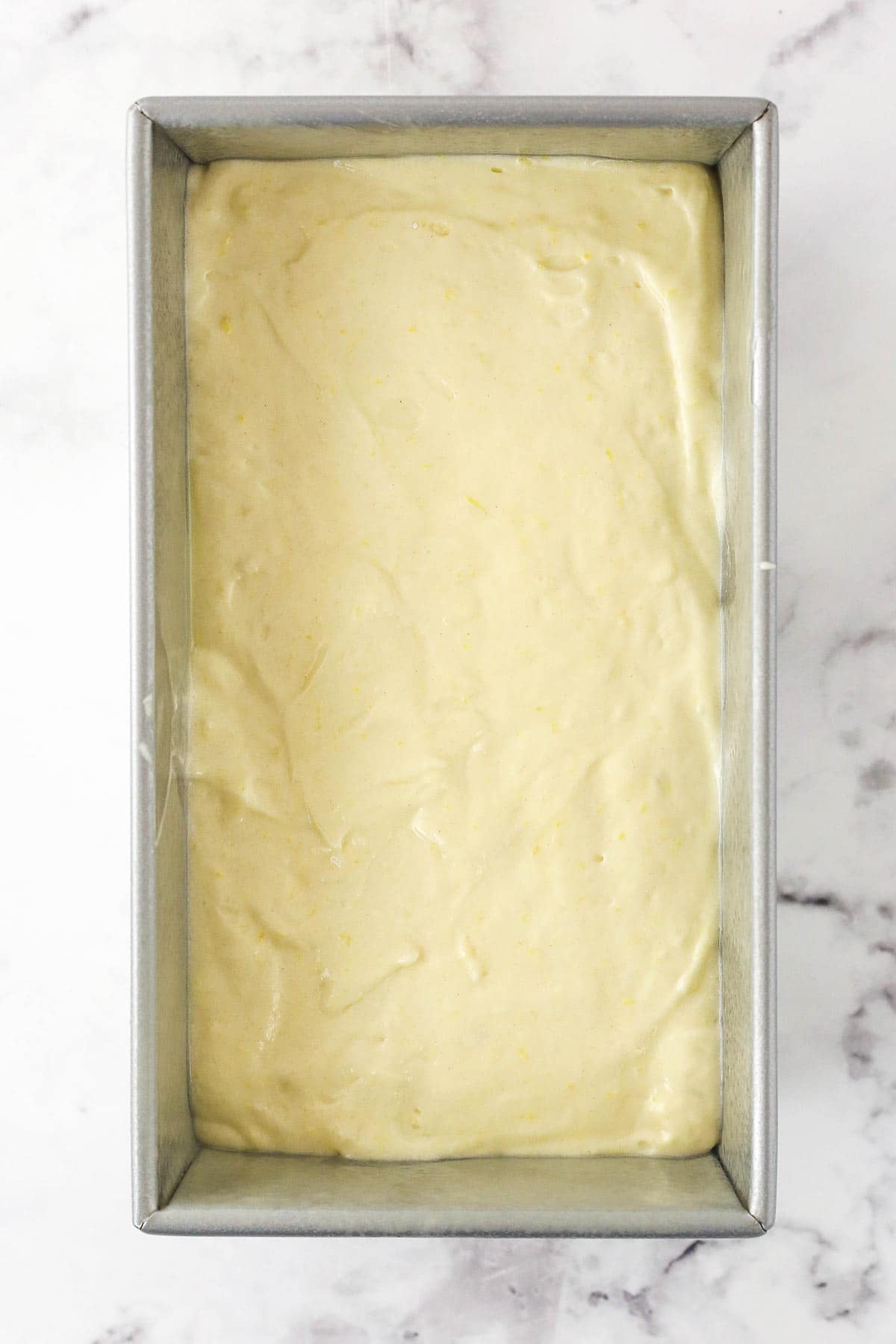 A loaf pan on a marble countertop with lemon breakfast cake batter inside of it