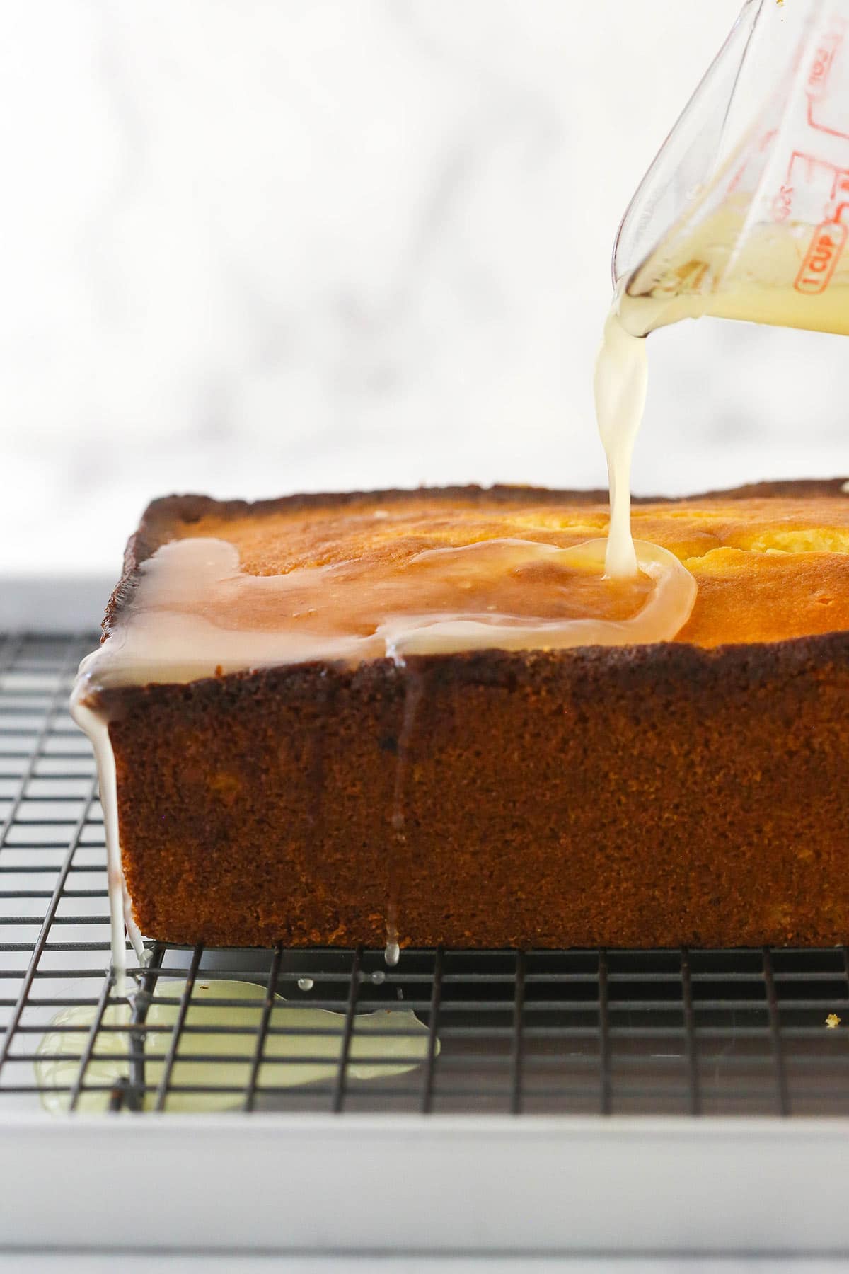 Lemon syrup being poured over the warm loaf cake while it's on a cooling rack with a baking sheet underneath it