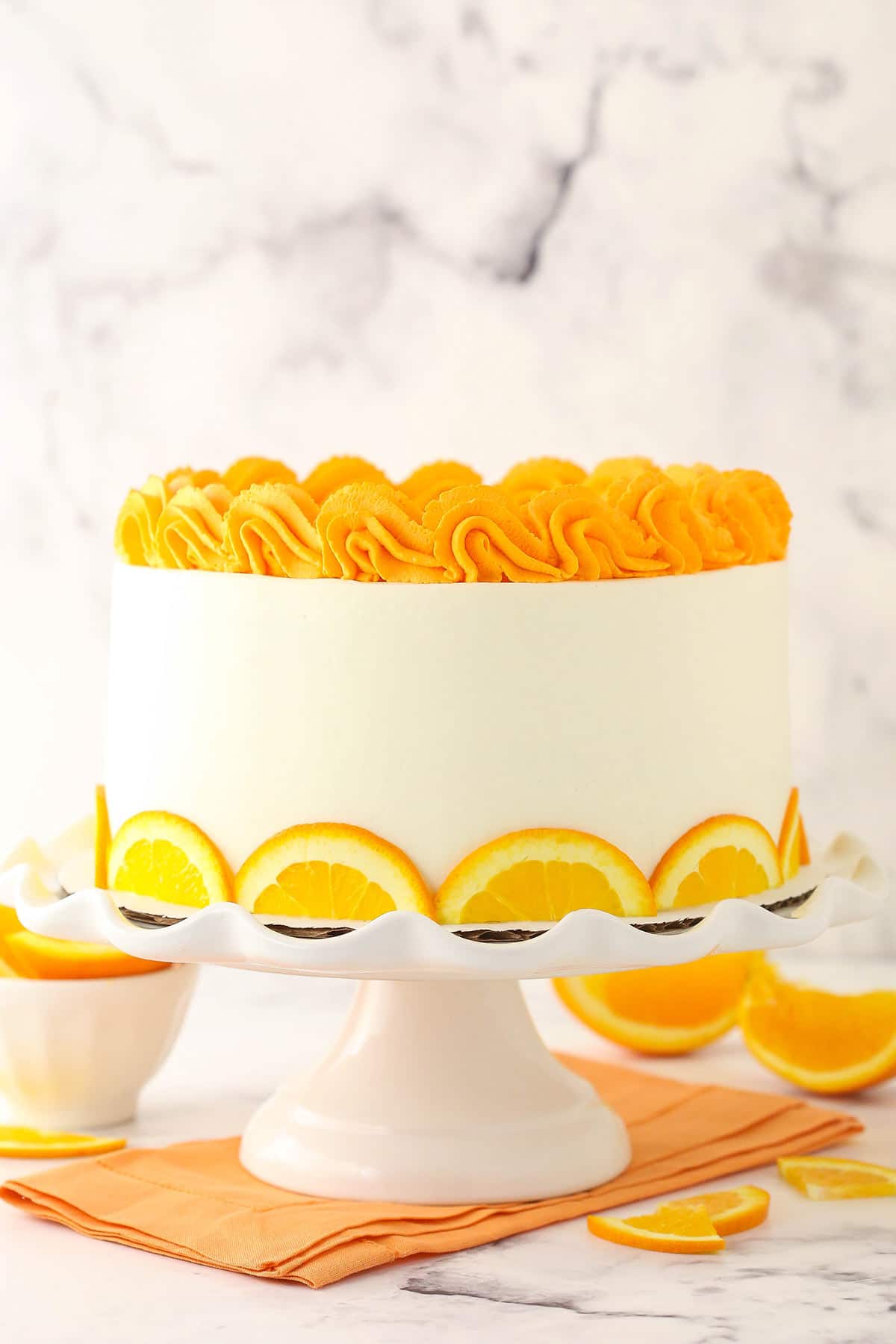 An orange layer cake on a tall white cake stand on top of an orange napkin