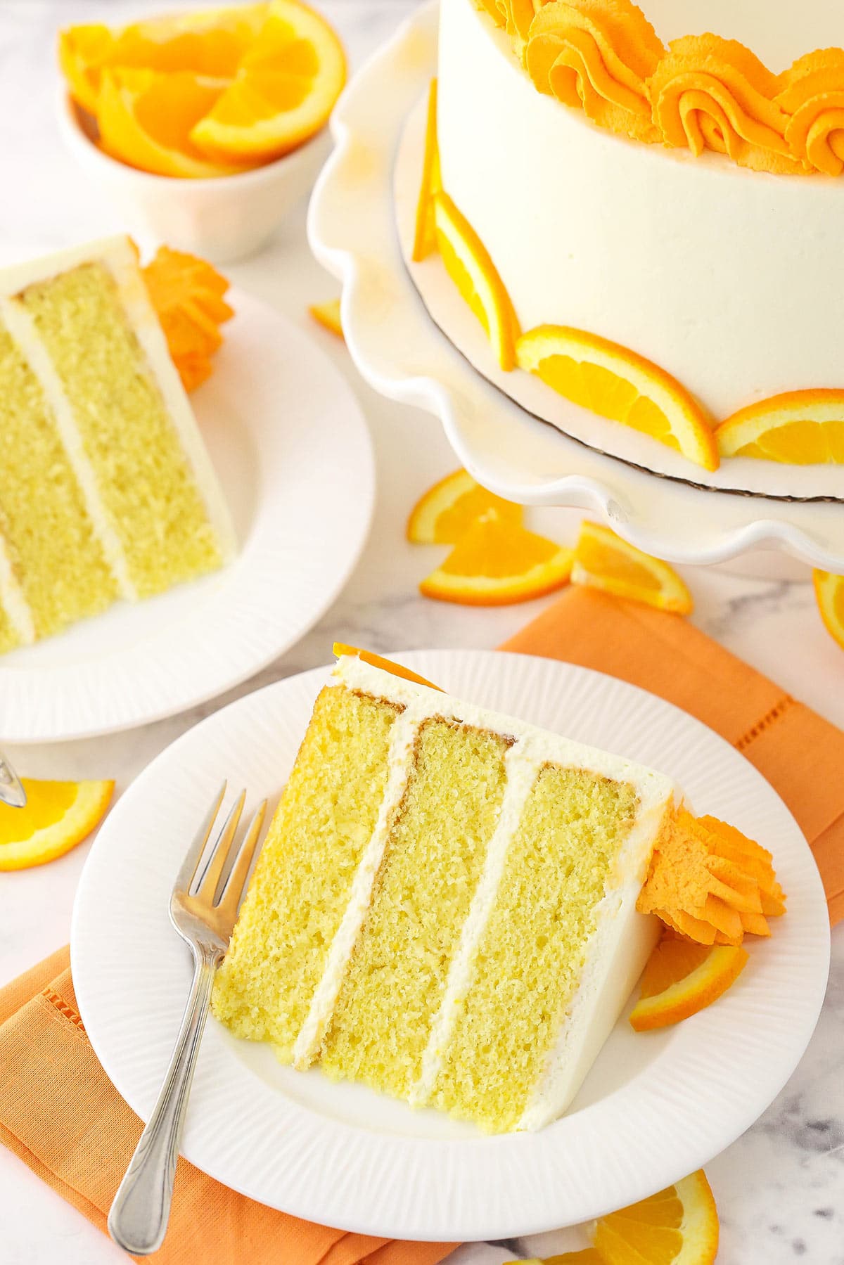 A slice of cake on a plate with a fork and a bowl of orange slices in the background