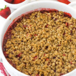 Baked strawberry rhubarb crisp in a round white baking dish with bowl of strawberries in background
