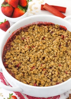 Baked strawberry rhubarb crisp in a round white baking dish with bowl of strawberries in background