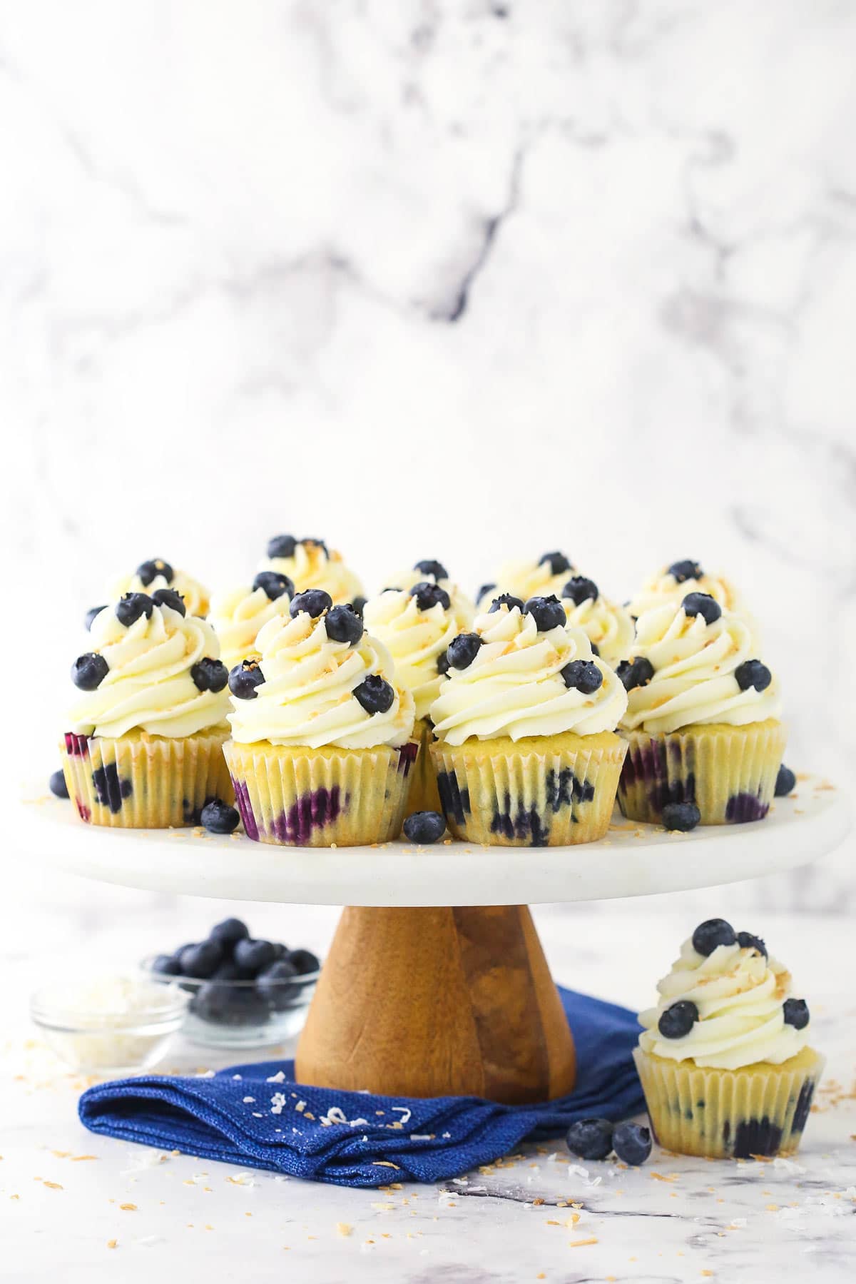 A batch of blueberry coconut cupcakes on a cake stand on top of a marble counter