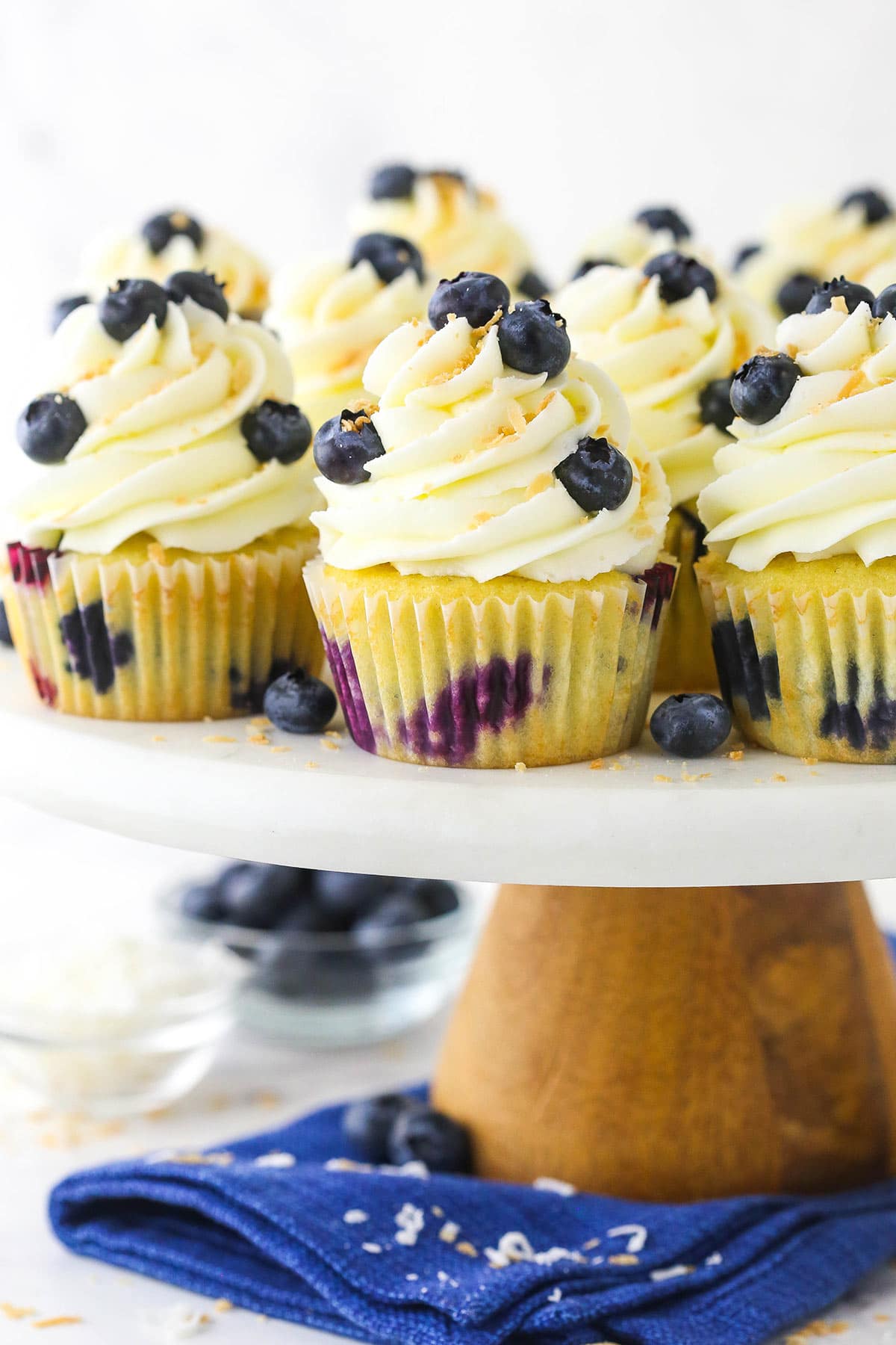 A cake stand holding a bunch of coconut cupcakes with a blue napkin underneath it