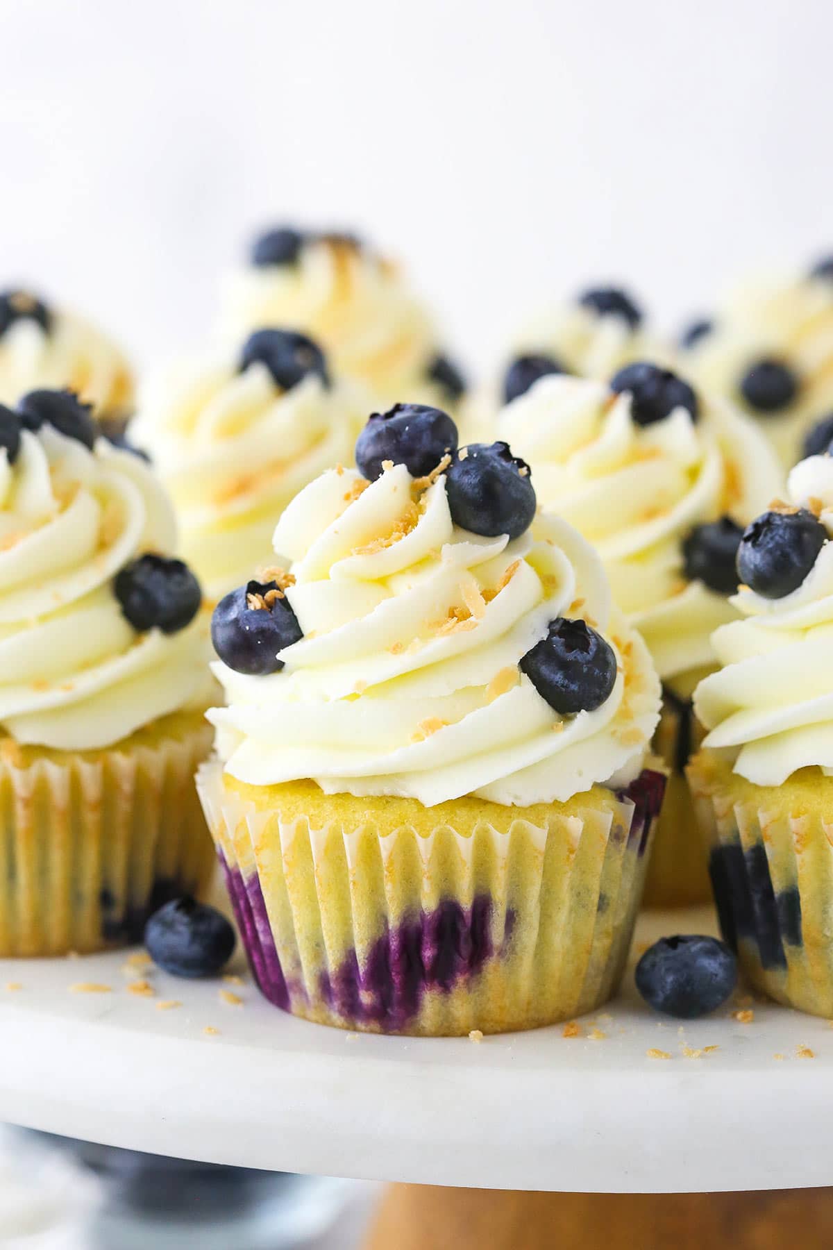 Easy Blueberry Coconut Cupcakes | Life Love & Sugar A batch of homemade cupcakes on a plastic cake stand with a wooden base