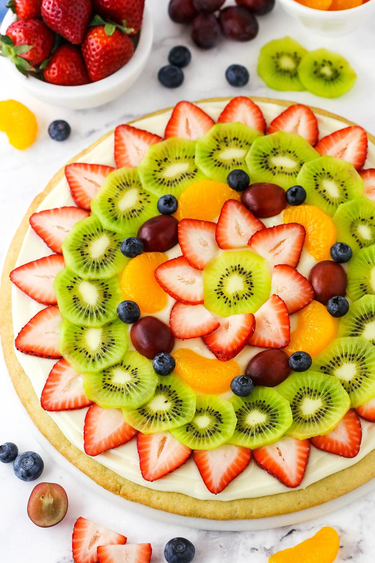 A sugar cookie fruit pizza on a kitchen countertop with assorted fruit surrounding it