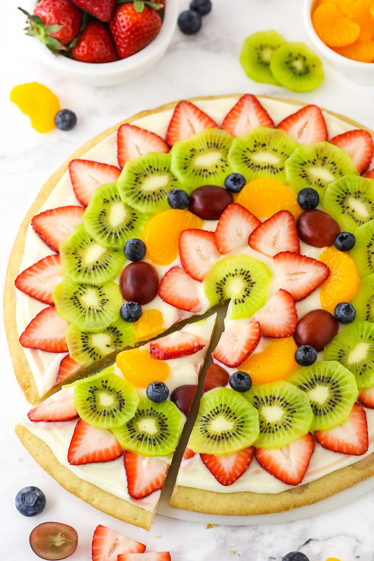 A homemade fruit pizza on a marble countertop with one slice cut out from the pizza