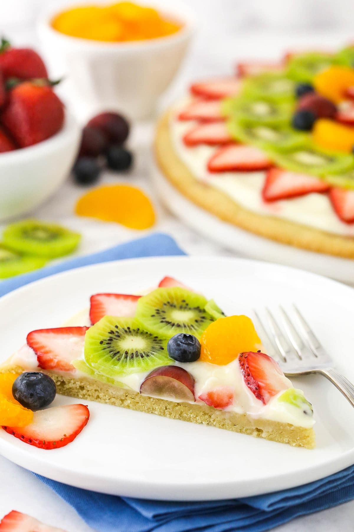A slice of fruit pizza on a plate with the remaining cookie pizza behind it