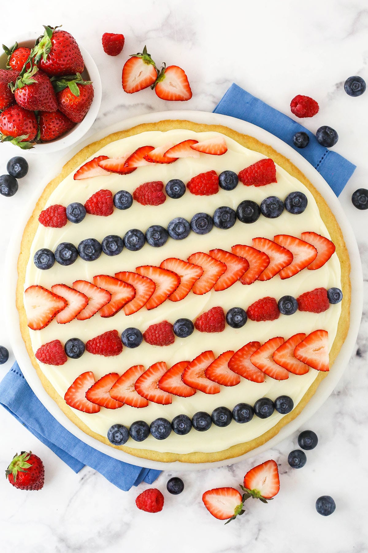 A large round sugar cookie base topped with cream cheese frosting, strawberries, raspberries and blueberries