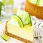 A slice of margarita cheesecake on a plate with the rest of the cheesecake on a cake stand behind it