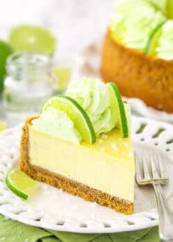 A slice of margarita cheesecake on a plate with the rest of the cheesecake on a cake stand behind it