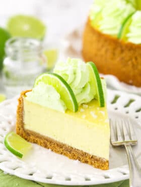 A slice of margarita cheesecake on a plate with the rest of the cheesecake on a cake stand behind it