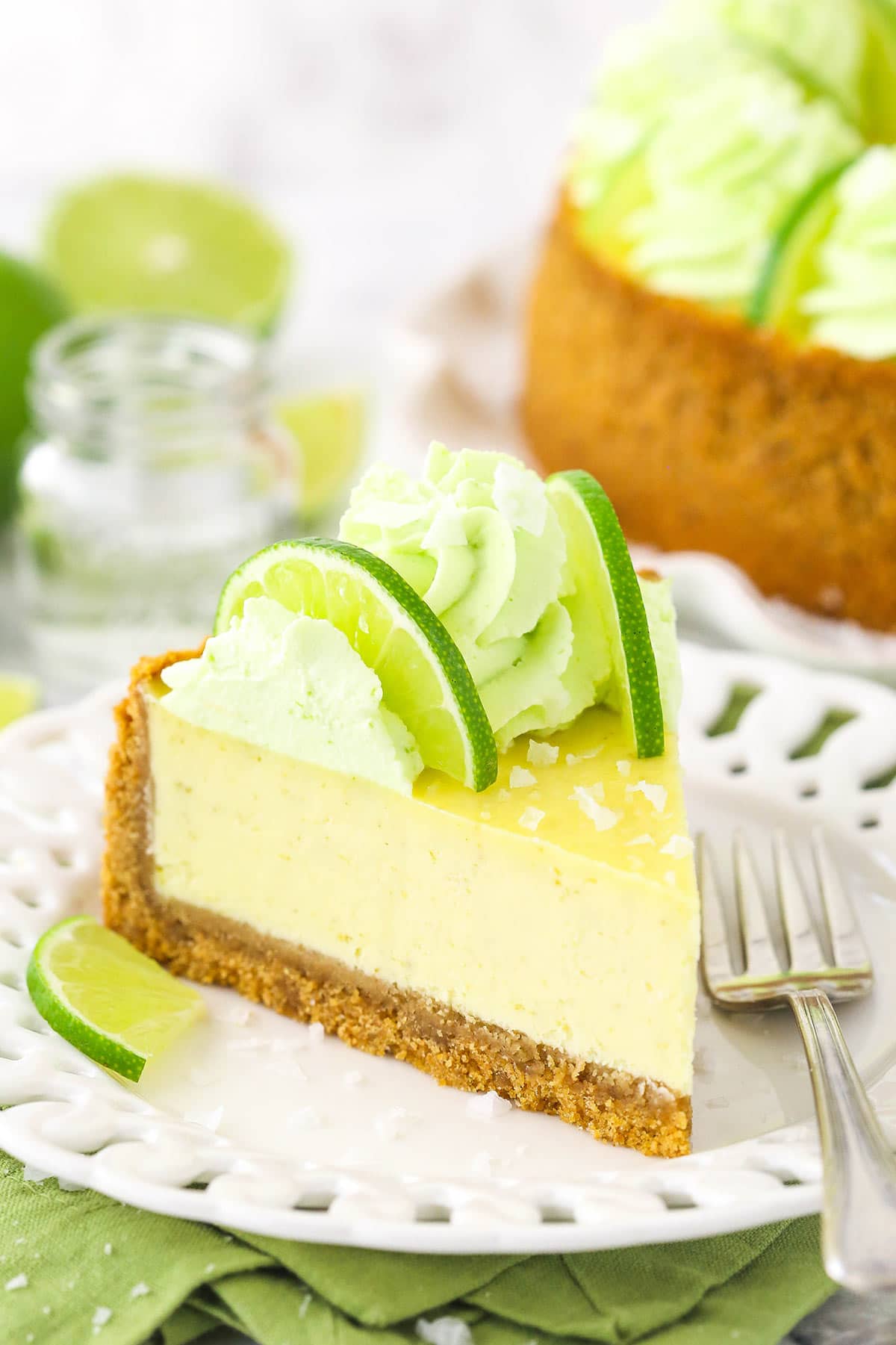 A slice of margarita cheesecake on a plate with the rest of the cheesecake on a cake stand behind it