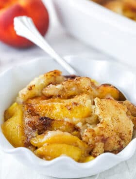 peach cobbler in white ruffle bowl with peach in background