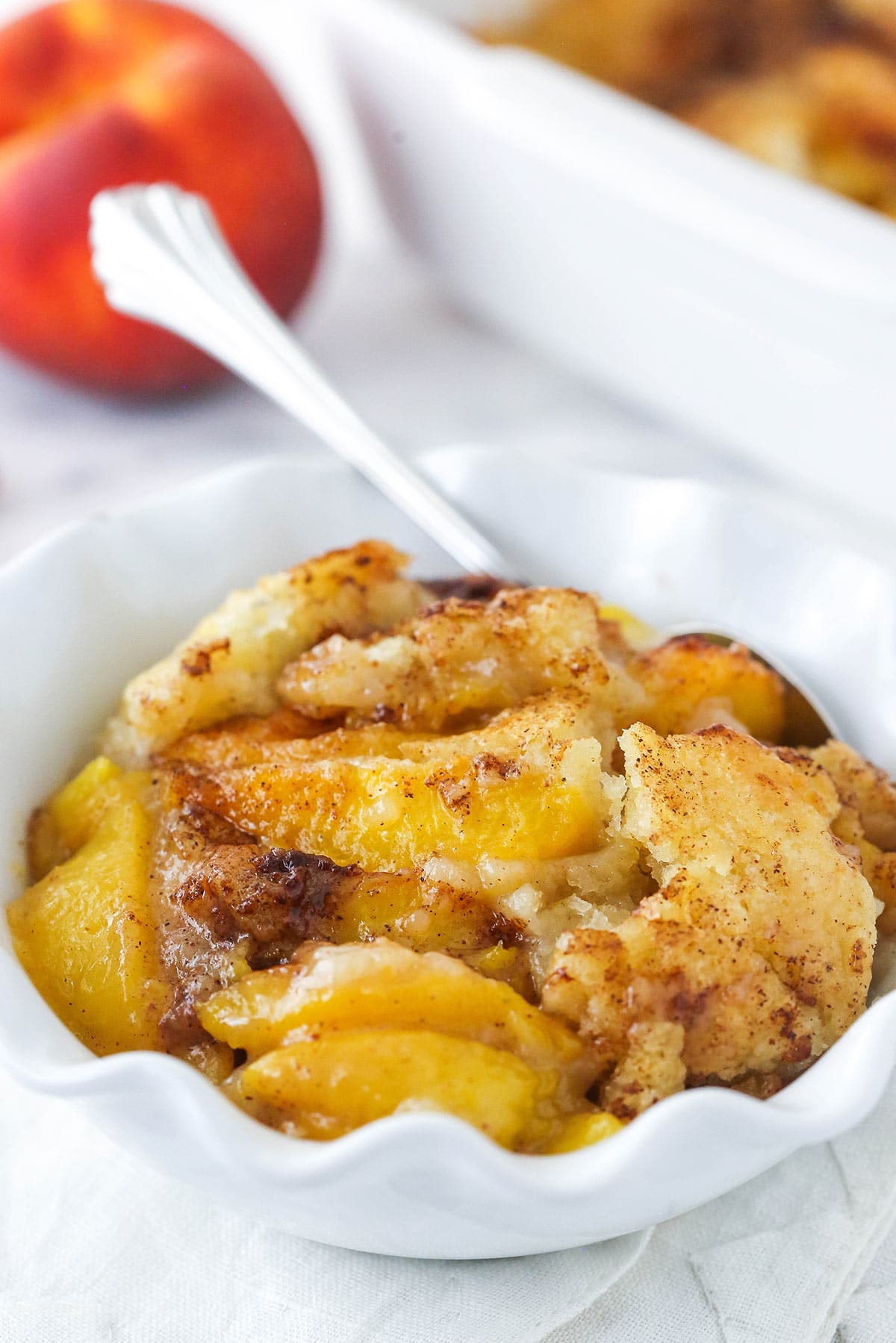 peach cobbler in white ruffle bowl with peach in background