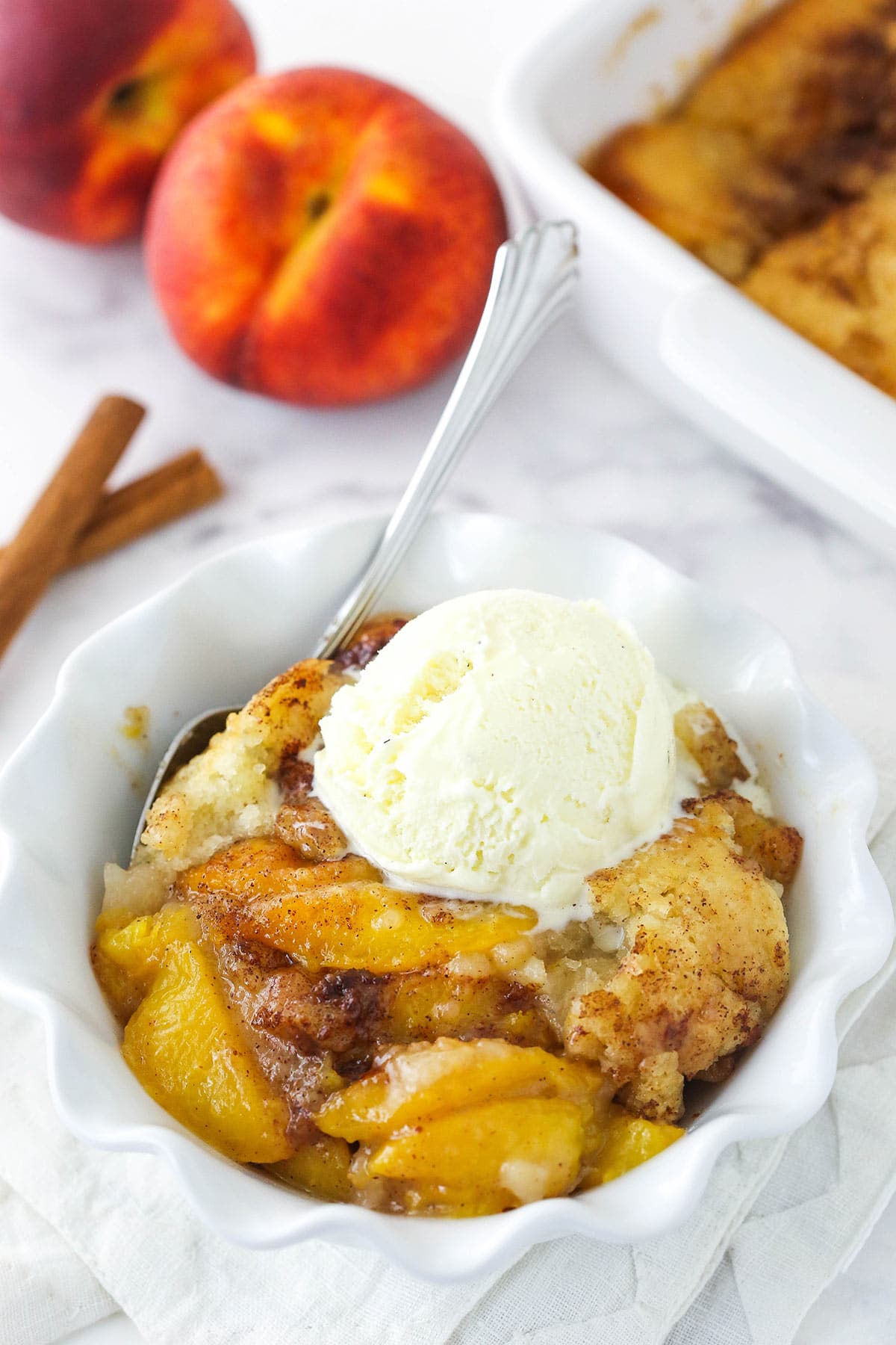peach cobbler in white bowl with scoop of ice cream on top and peached in background