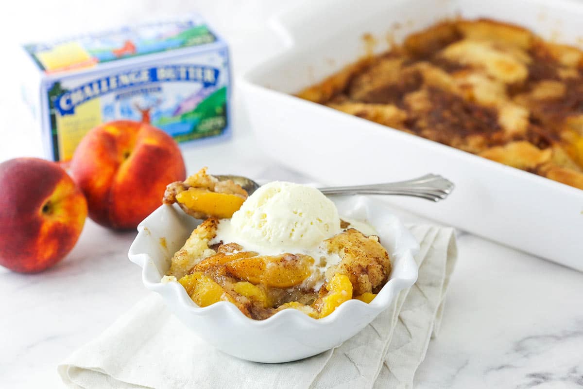 peach cobbler in white bowl with bite scooped out and casserole dish, butter and peaches in the background