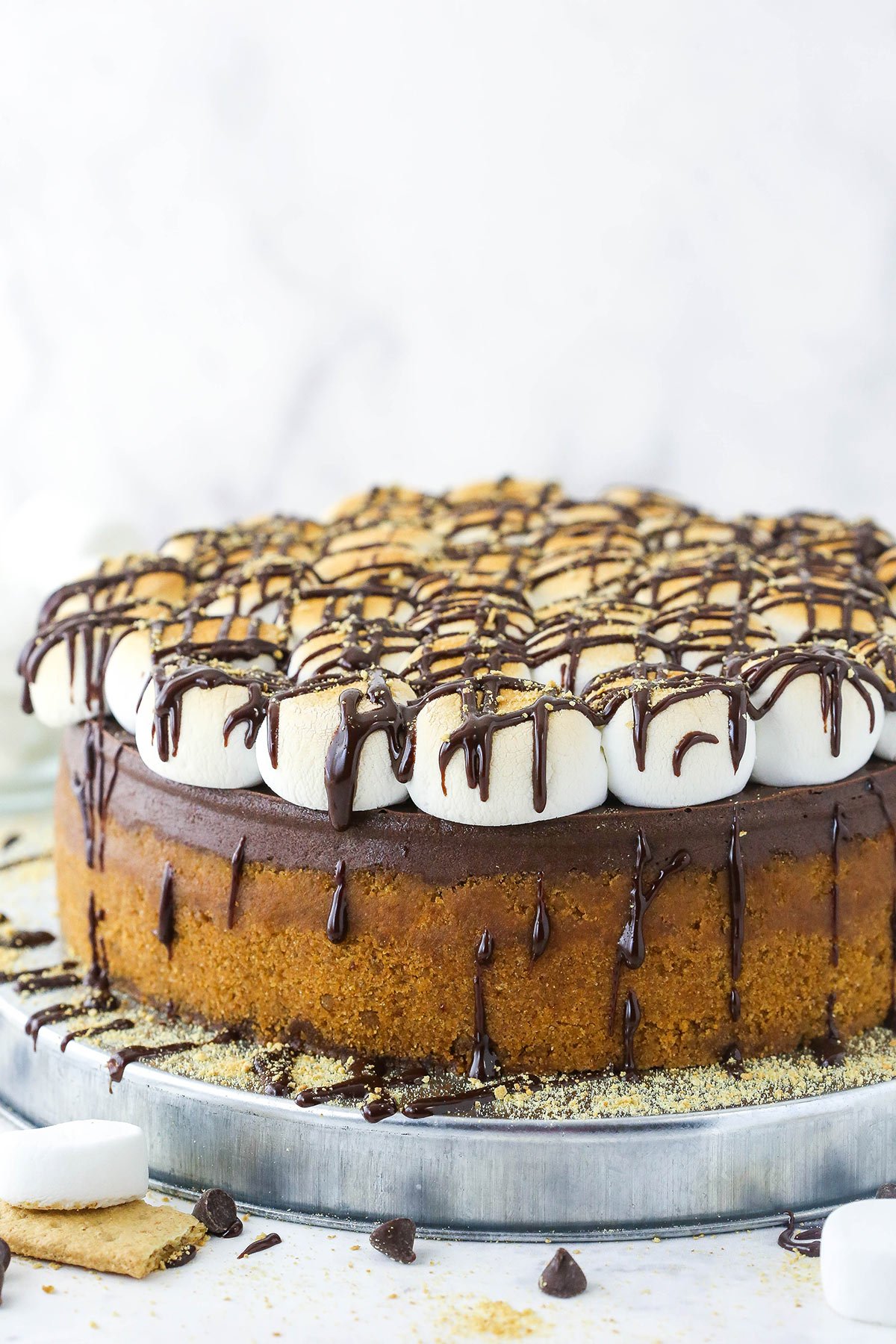 A smores cheesecake on a metal cake stand with marshmallows, chocolate chips and graham crackers on the counter next to it