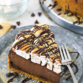 A piece of graham cracker, chocolate and marshmallow cheesecake on a plate with a bowl of marshmallows behind it