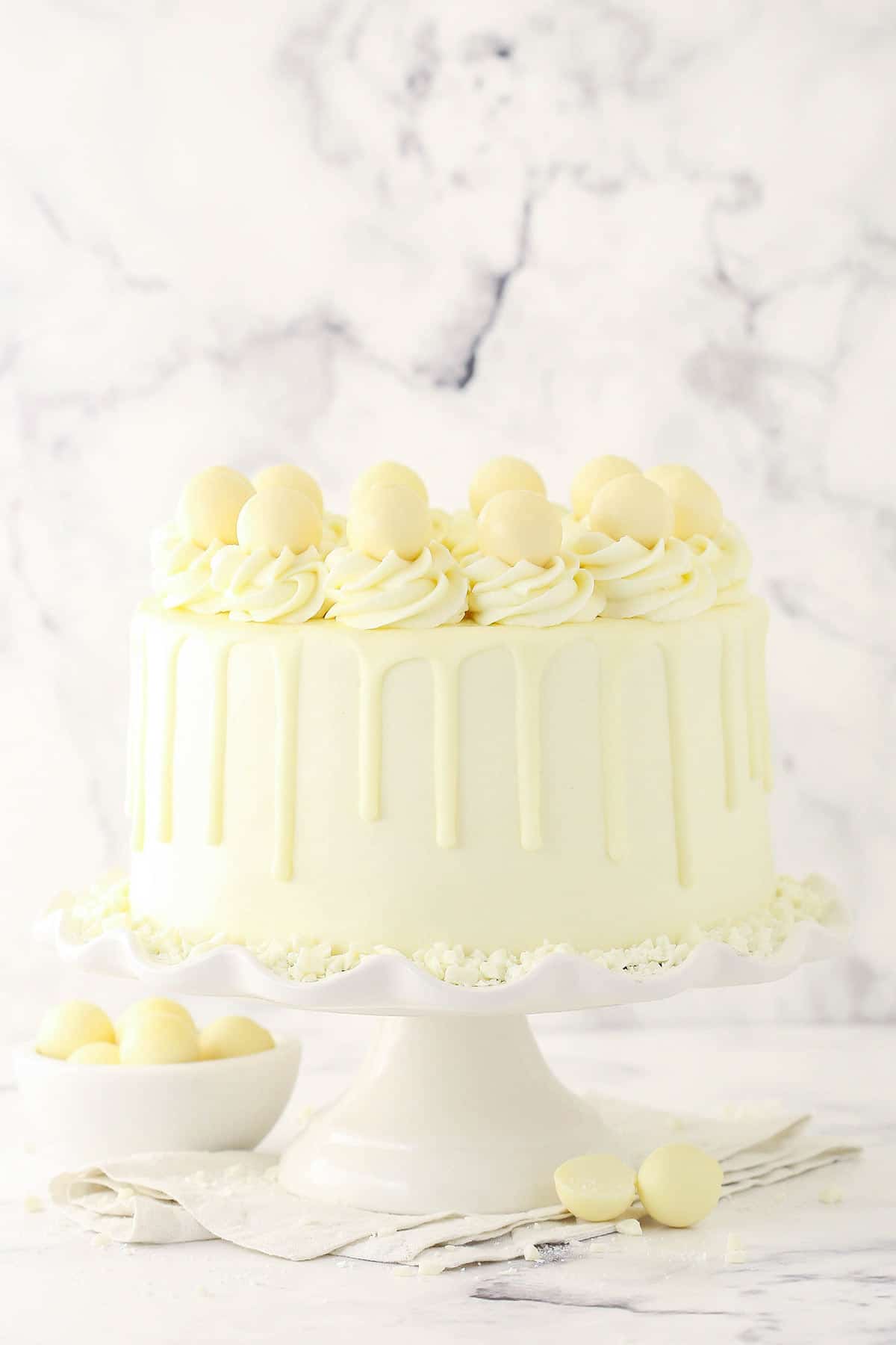 A cake stand holding a white chocolate layer cake with a white cloth napkin underneath it