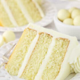 A piece of white chocolate cake on a plate with a bowl of chocolate truffles and a second slice of cake in the background