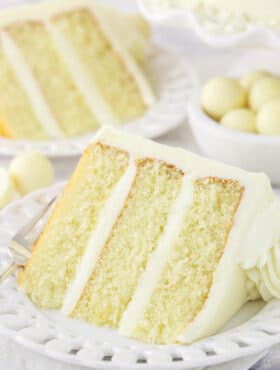 A piece of white chocolate cake on a plate with a bowl of chocolate truffles and a second slice of cake in the background