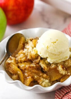 A close-up shot of apple cobbler in a bowl with two apples in the background