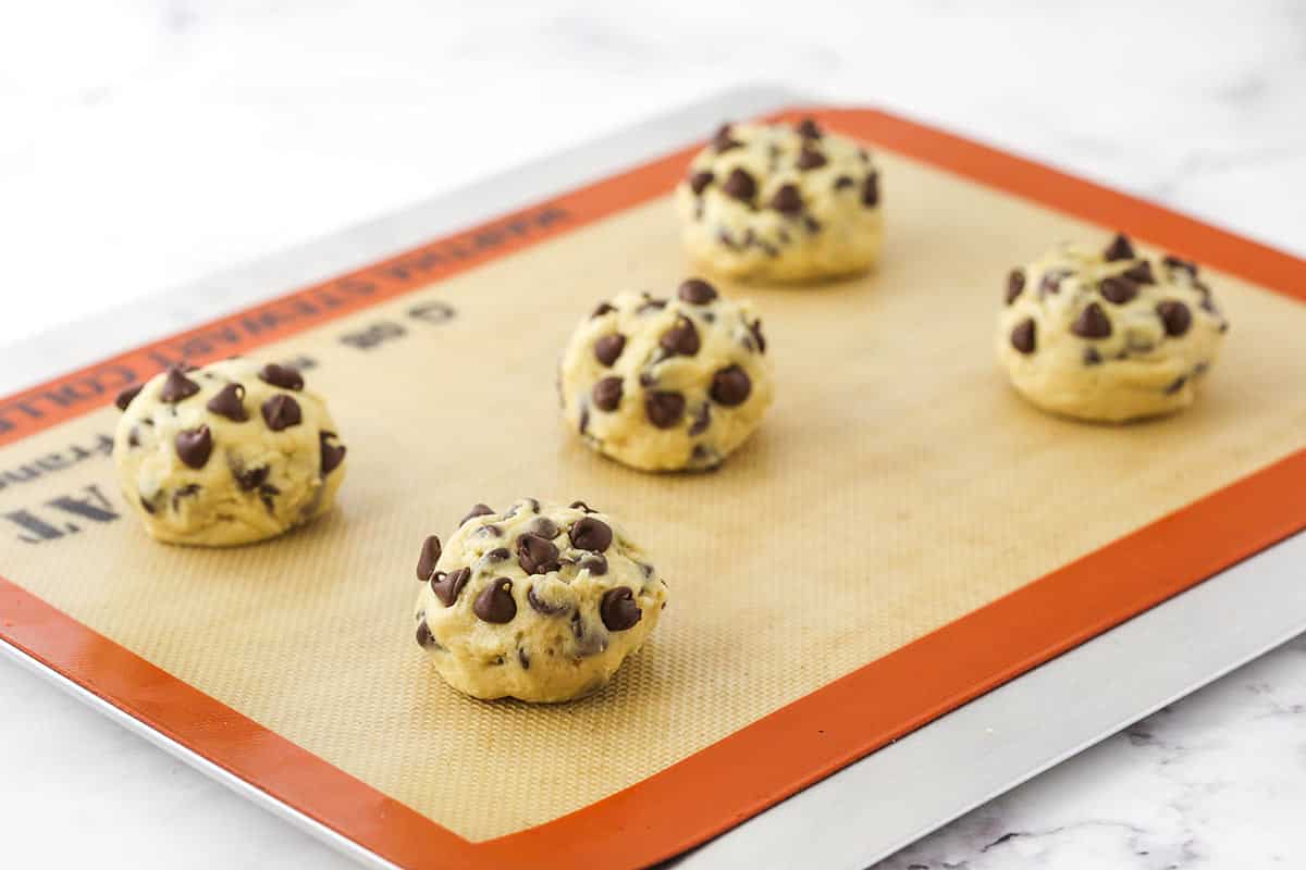 Five balls of cookie dough on a baking sheet lined with a Silpat baking mat.