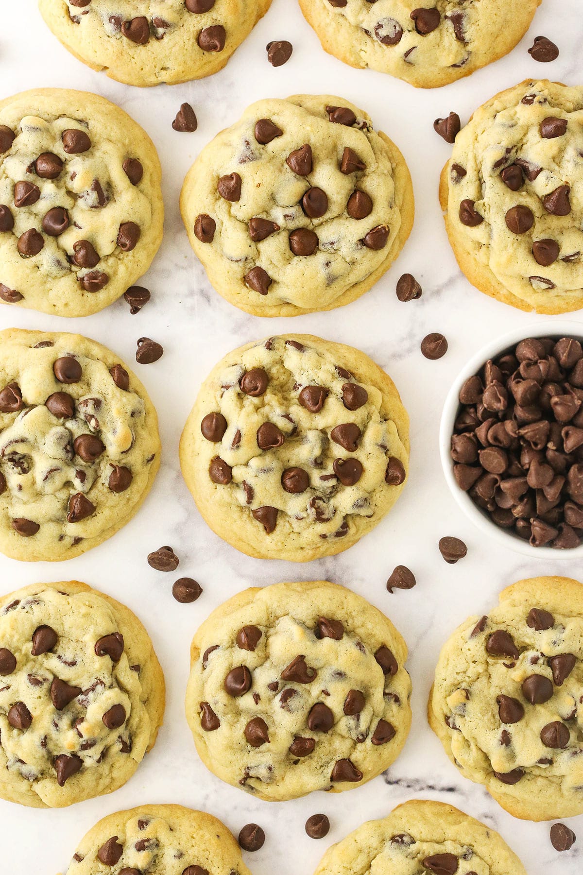 A bunch of thick and chewy chocolate chip cookies on a marble surface with a small bowl of chocolate chips