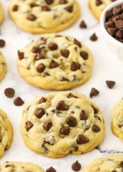 Homemade chocolate chip cookies lined up on a kitchen countertop with some loose chocolate chips