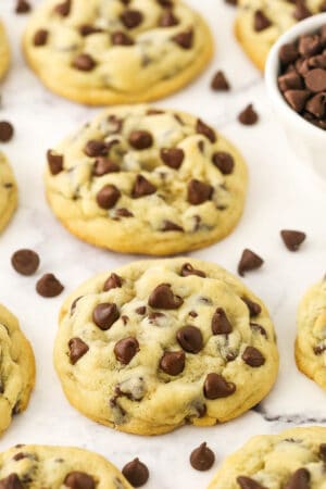 Homemade chocolate chip cookies lined up on a kitchen countertop with some loose chocolate chips